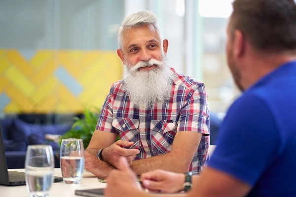 Bearded man sitting at table speaking to someone faced away from the camera