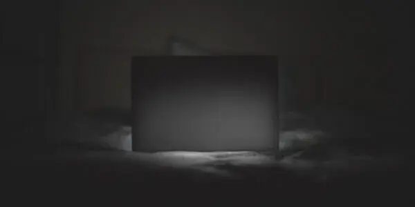 Sinister image of laptop on bed illuminated in dark room