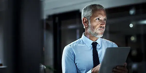 Middle-aged business professional standing in dark office environment holding digital tablet device